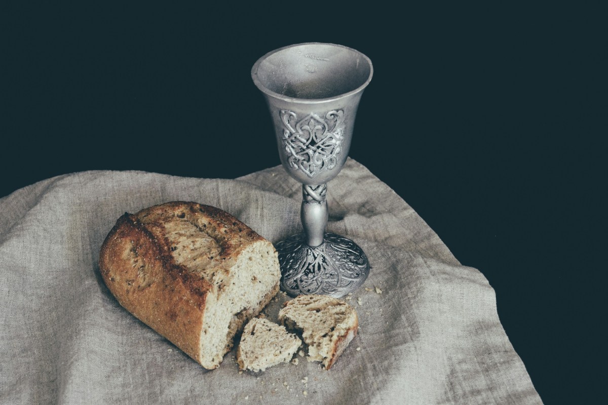 A silver chalice and a loaf of bread with broken pieces rest on a linen cloth against a dark background.